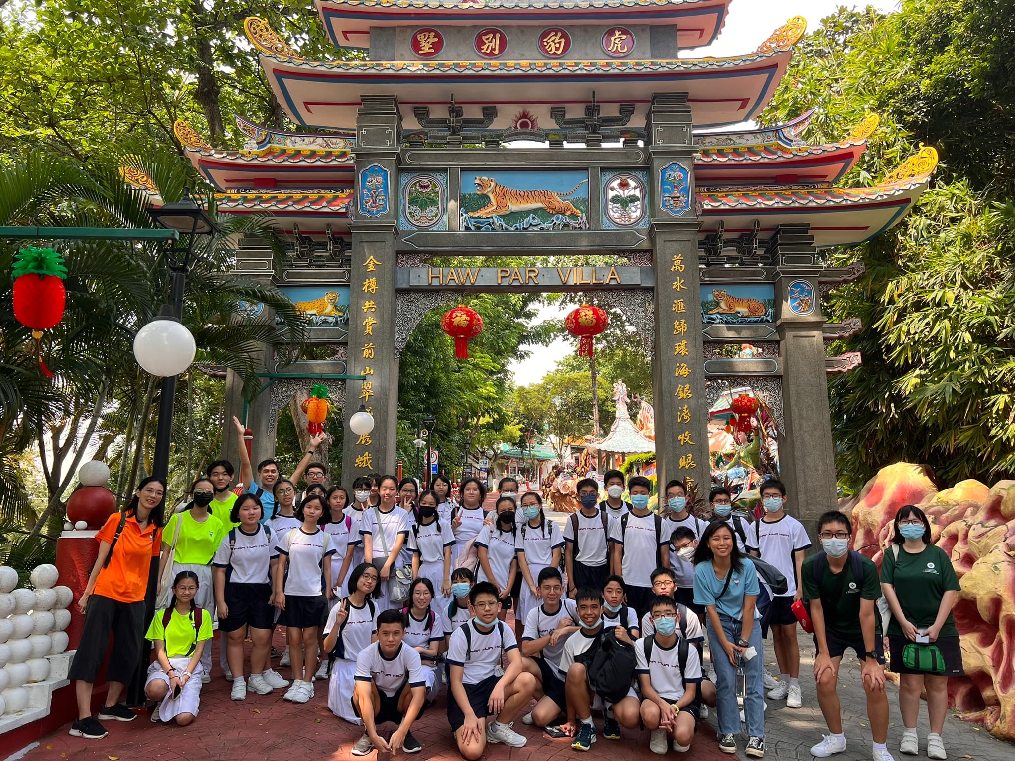 A large group of students in white uniforms pose under the ornate entrance gate of Haw Par Villa in Singapore.